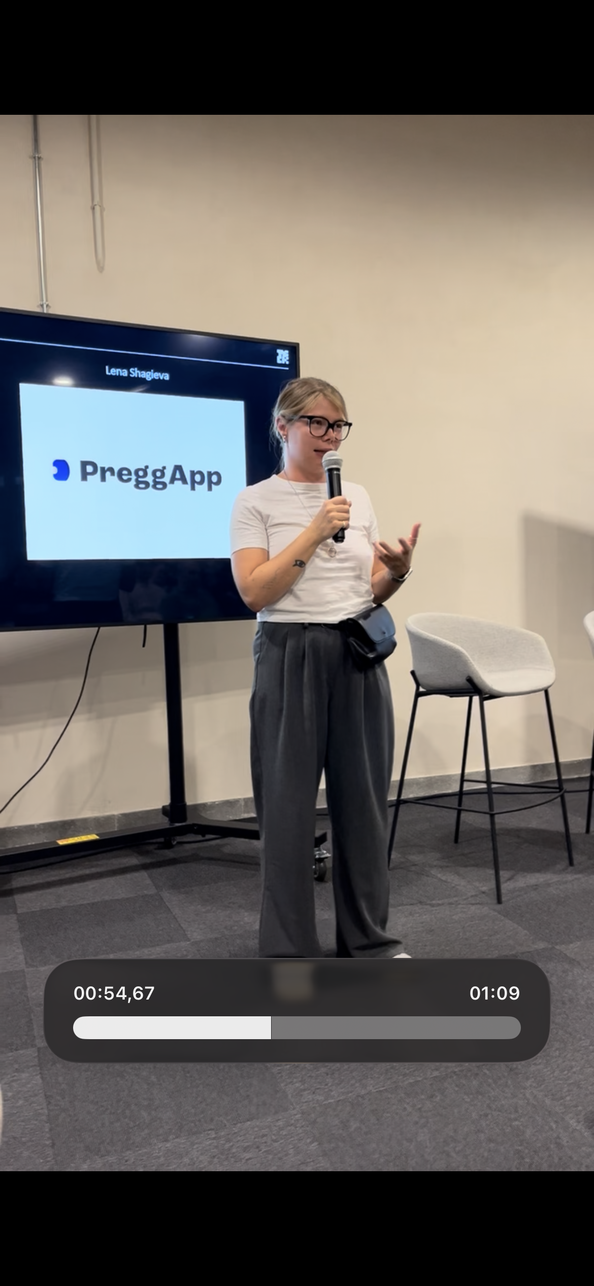 A woman with blonde hair and glasses speaking into a microphone during a presentation, standing in front of a screen displaying the logo for PreggApp.