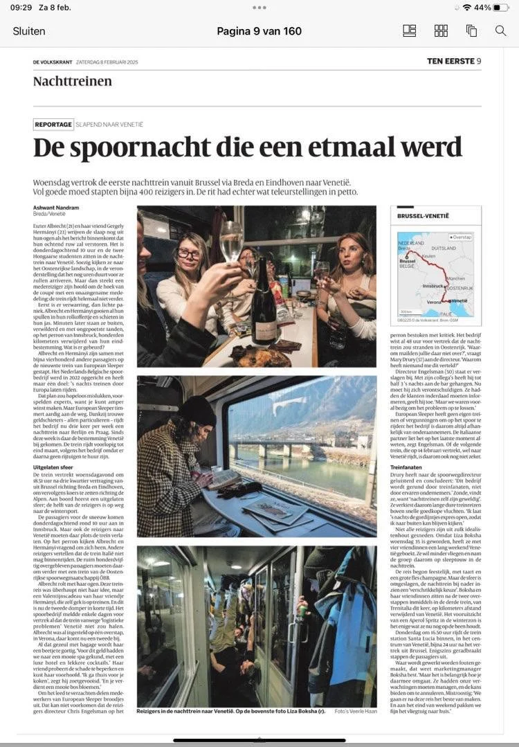 Three women sitting at a dinner table, raising glasses for a toast, with a bottle of champagne and a wine glass. One woman is wearing glasses and smiling, while the others are looking at her. The setting appears to be a restaurant or a social gathering.