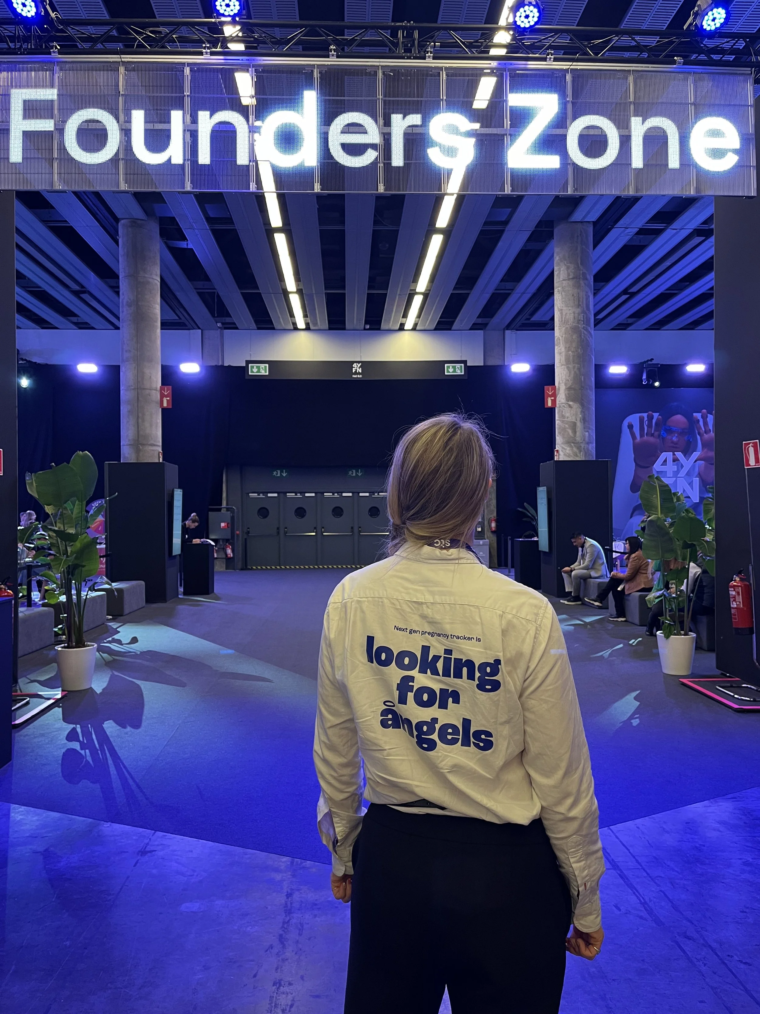 Person standing in front of a large illuminated sign that reads 'Founders Zone' inside a modern, well-lit area with chairs, potted plants, and a black backdrop.