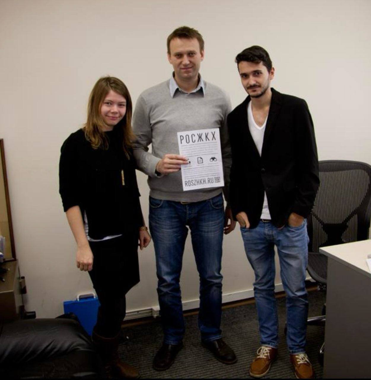 Three young adults standing indoors, the man in the middle holding a flyer with Cyrillic text and a website link.