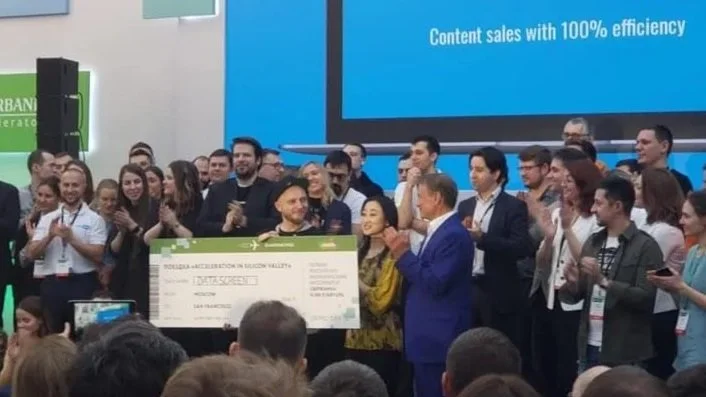 Group of people at an award ceremony, with a man in a blue suit presenting a large check to a woman, surrounded by applause and smiling attendees.