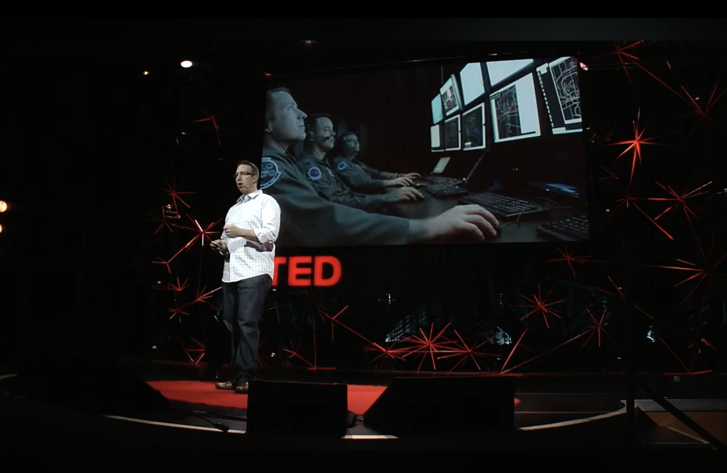 A man giving a talk at a TED conference, standing on stage with a large screen behind him showing a group of people working on computers, with the TED logo illuminated in red.