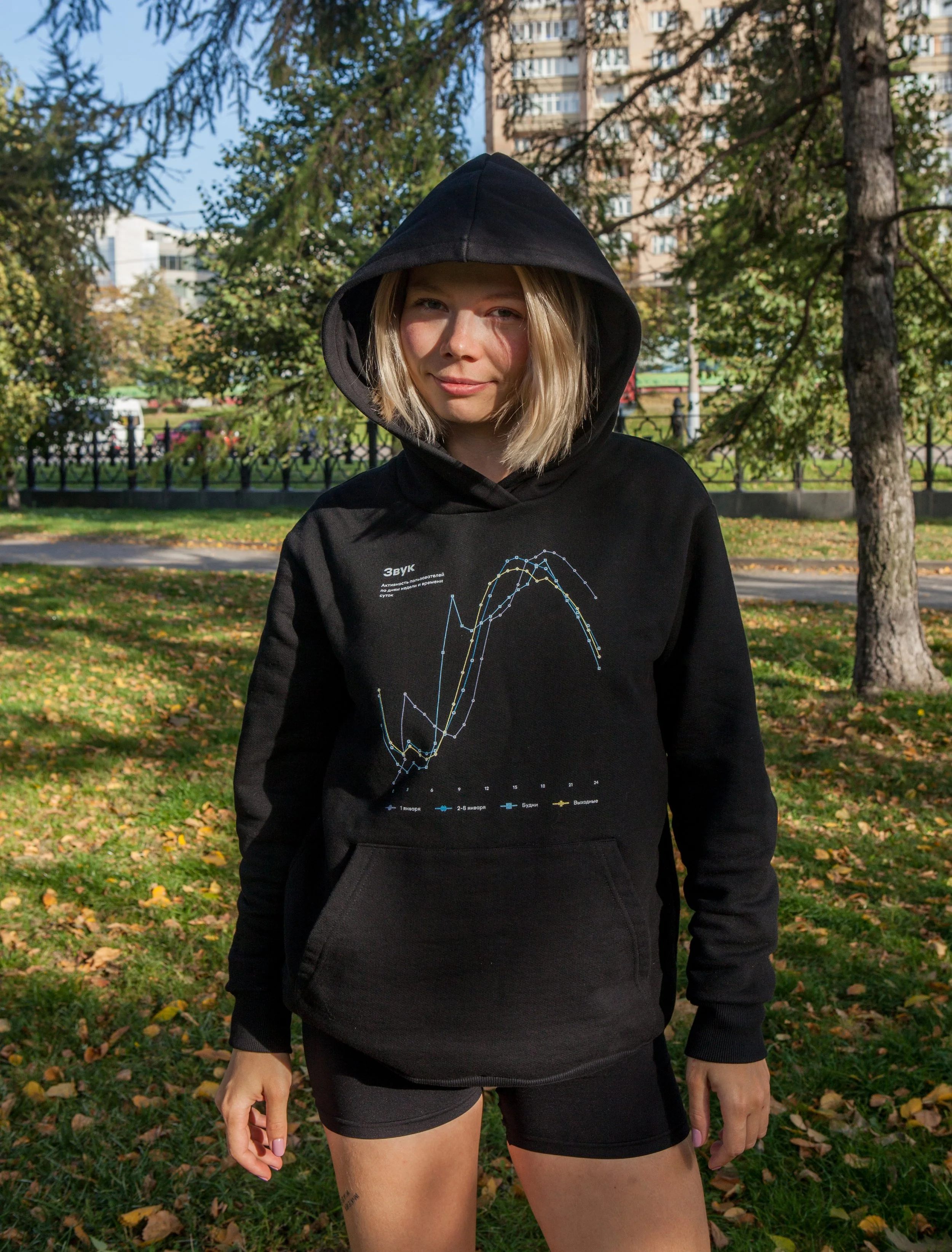 A young woman with blonde hair wearing a black hoodie with a graph printed on the front, standing outdoors in a park with trees and buildings in the background.