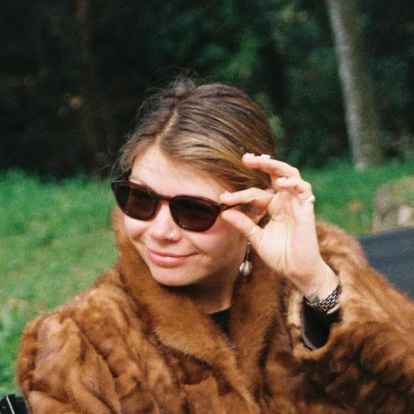 A woman wearing sunglasses and a fur coat outdoors, with trees and grass in the background.