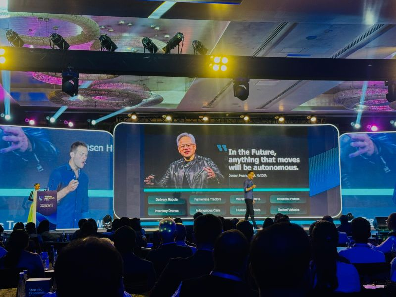 A technology conference with a large stage featuring two presenters and a large screen. The screen displays a quote about autonomous technology from Jensen Huang, CEO of NVIDIA. The presenters are speaking to an audience seated in rows.