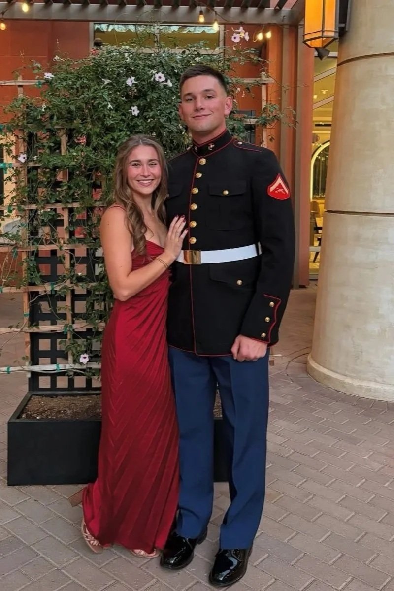 A young woman in a red evening dress and a young man in a Marine dress uniform are standing together, smiling, in an outdoor setting with plants and decorative architecture.