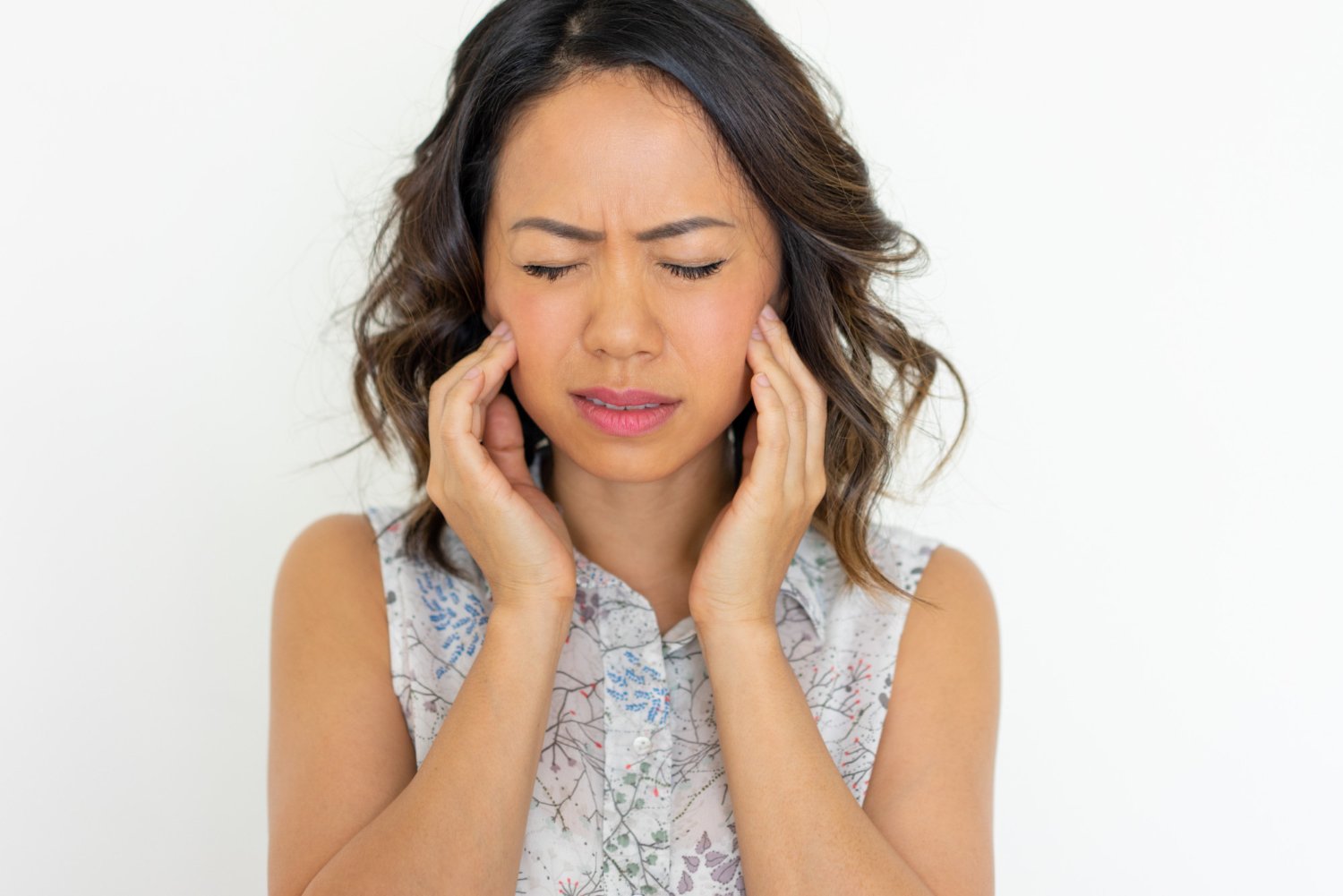 A woman with shoulder-length brown hair holding her head with hands, eyes closed, appearing to have a headache or ear pain, against a plain white background.