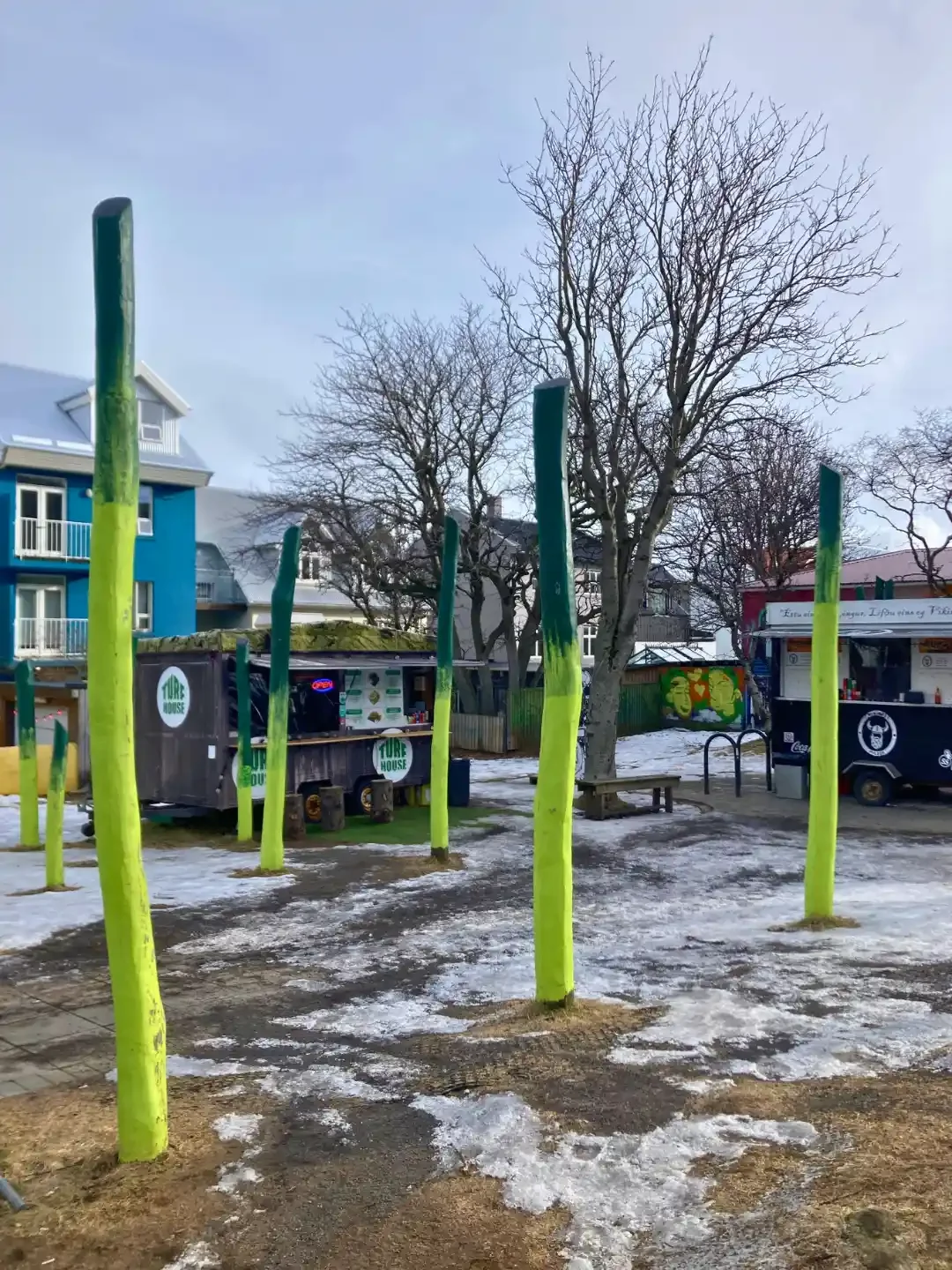 Neon green sculptural poles stand in snowy downtown Reykjavik beside cafés and bare winter trees.