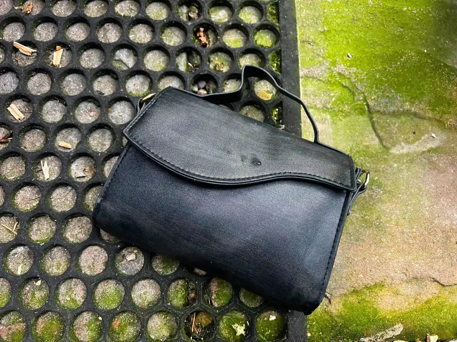 Black thrift store handbag on concrete with green moss, and black rug with holes in.