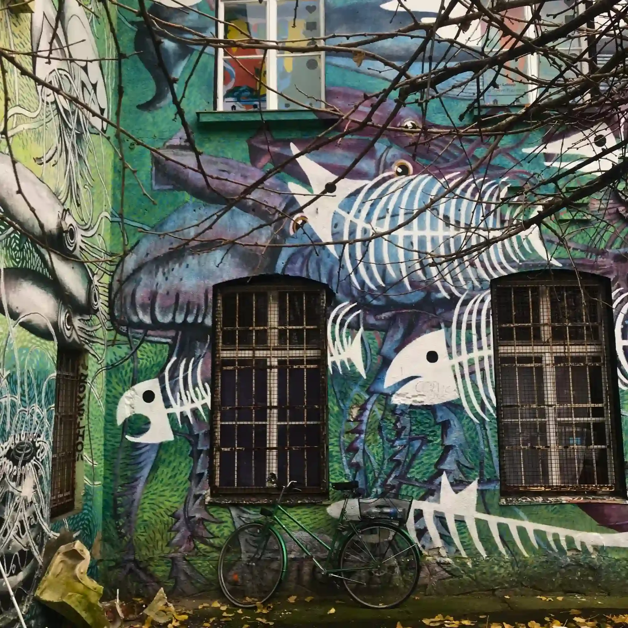 Colourful fish mural covers a building wall in Ljubljana’s Metelkova district, with bicycles and bare branches below.