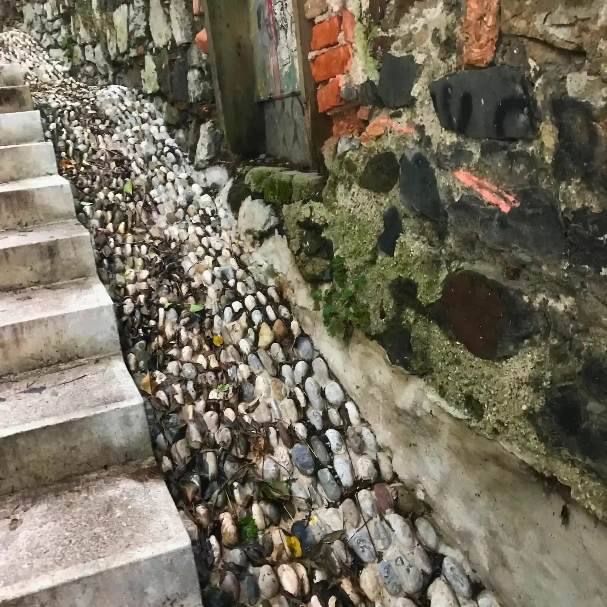 Stone wall with exposed brick, concrete steps, and a pebble-lined drainage path in an urban Slovenian alley.