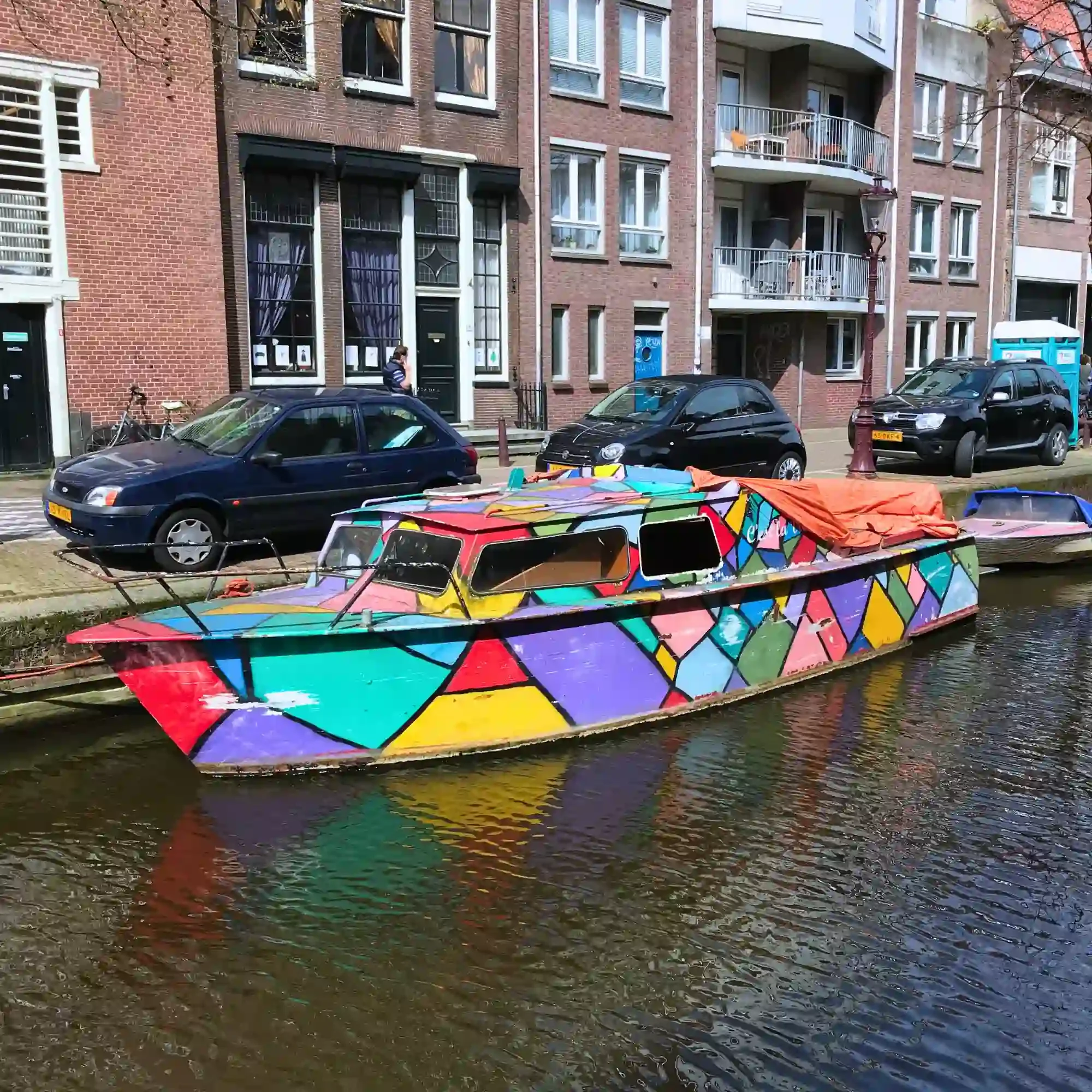 Colourful geometric canal boat moored beside Amsterdam apartments on a sunny city canal.