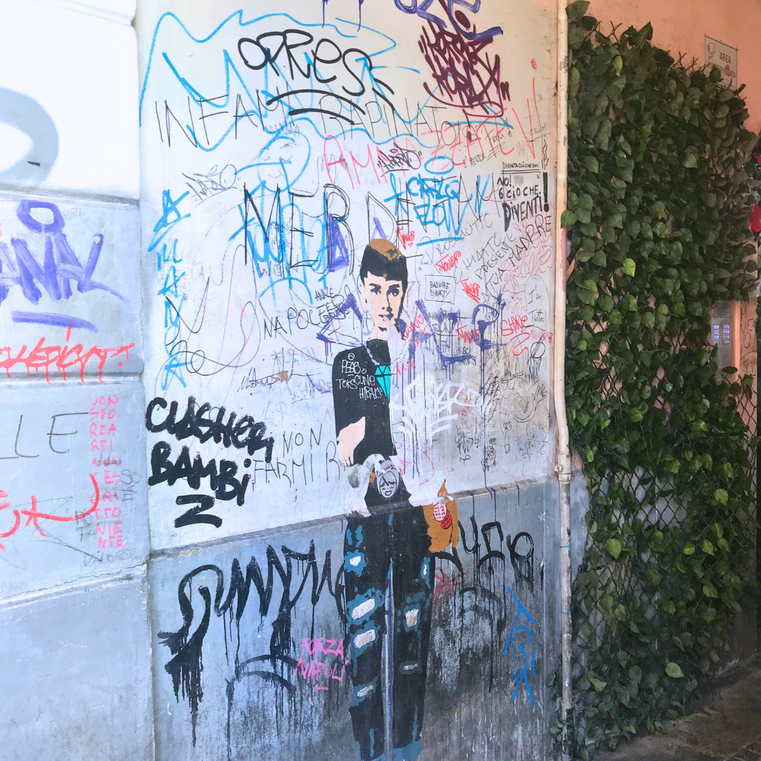 Woman painted on an urban wall next to urban graffiti and greenery.
