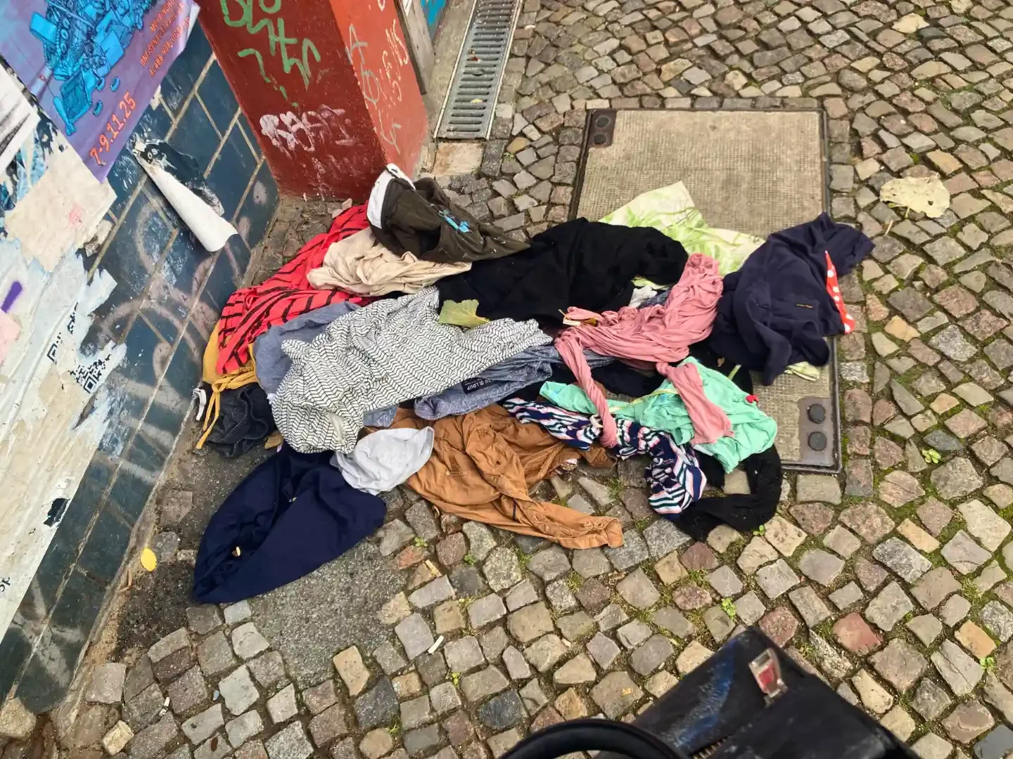 Discarded clothing and textile waste dumped on a cobbled city street beside a graffiti-covered wall and drain cover.