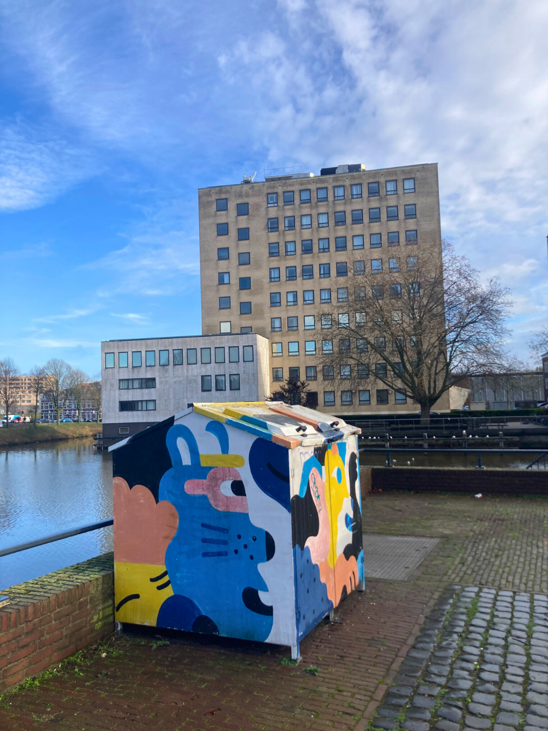 Colourful painted utility box beside a riverside walkway in an urban area with modern buildings and cobbled pavement.