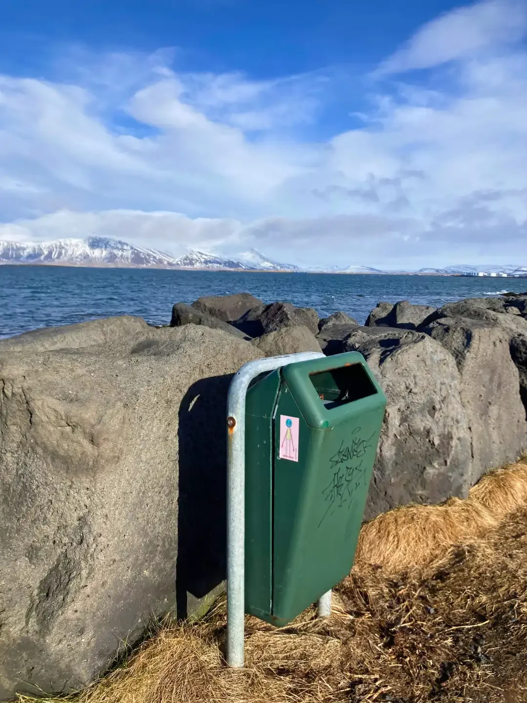 a green litter bin by grey rocks in front of the ocean with mountains in the background.