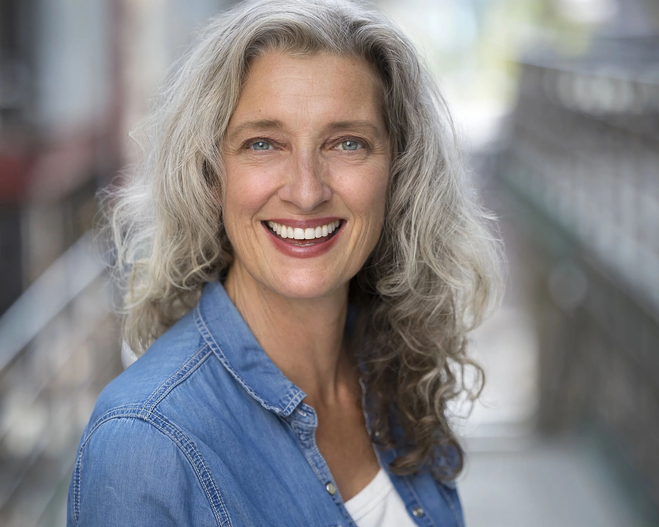 A close-up of a middle-aged woman with gray curly hair, smiling, wearing a blue denim jacket indoors.