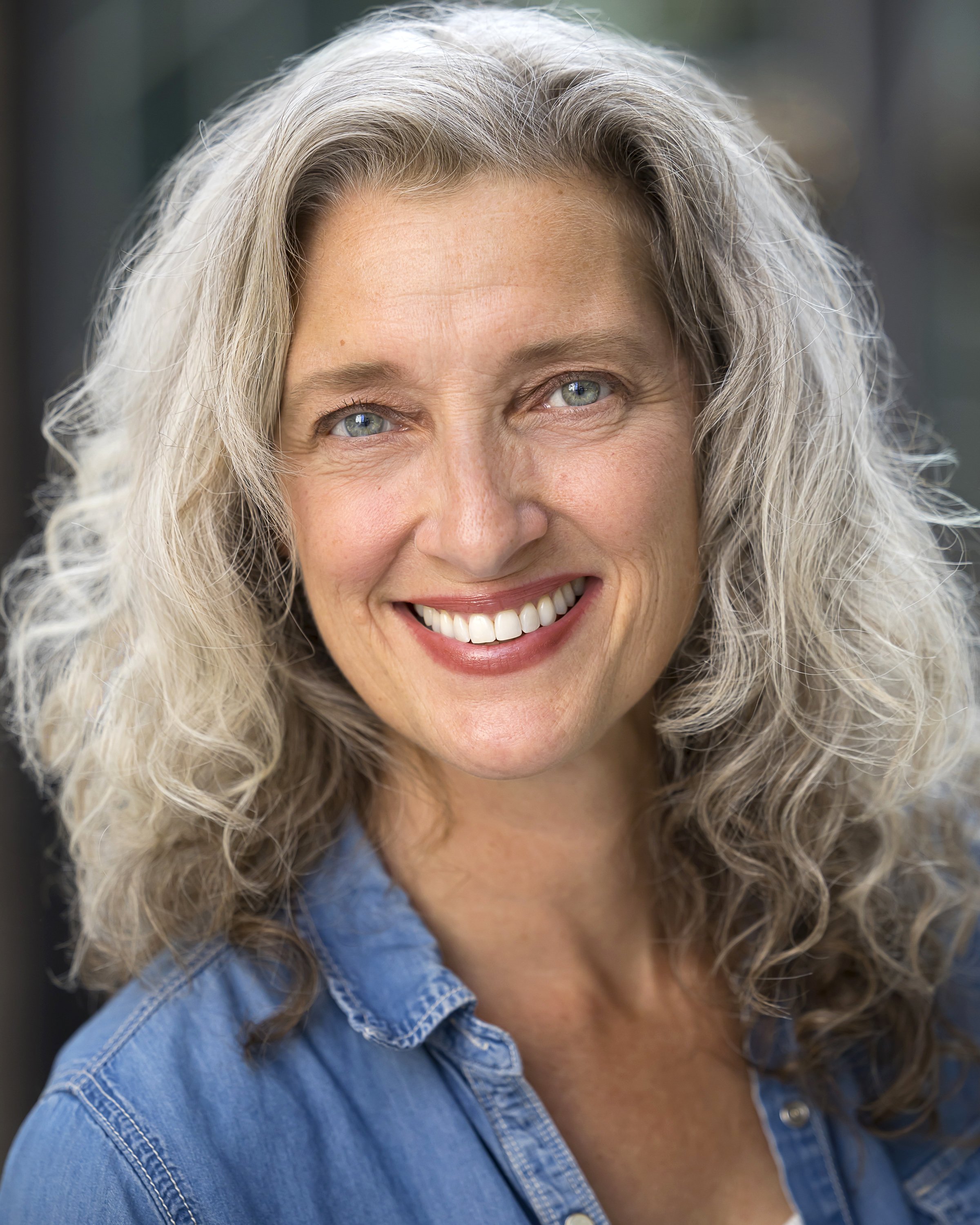 Close-up portrait of a woman with curly gray hair, blue eyes, smiling, wearing a denim shirt.