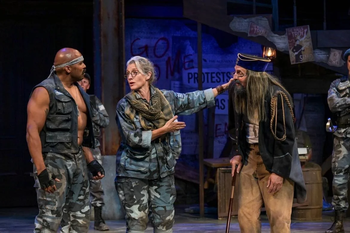 A stage scene with four actors: a muscular man in camouflage pants and a sleeveless vest, a woman in camouflage military outfit with glasses, an older man with a beard wearing a navy hat and blazer, and another man in camouflage military uniform in the background. The woman is touching the older man's forehead as if in a confrontation or conversation, with a backdrop of protest signs and posters.