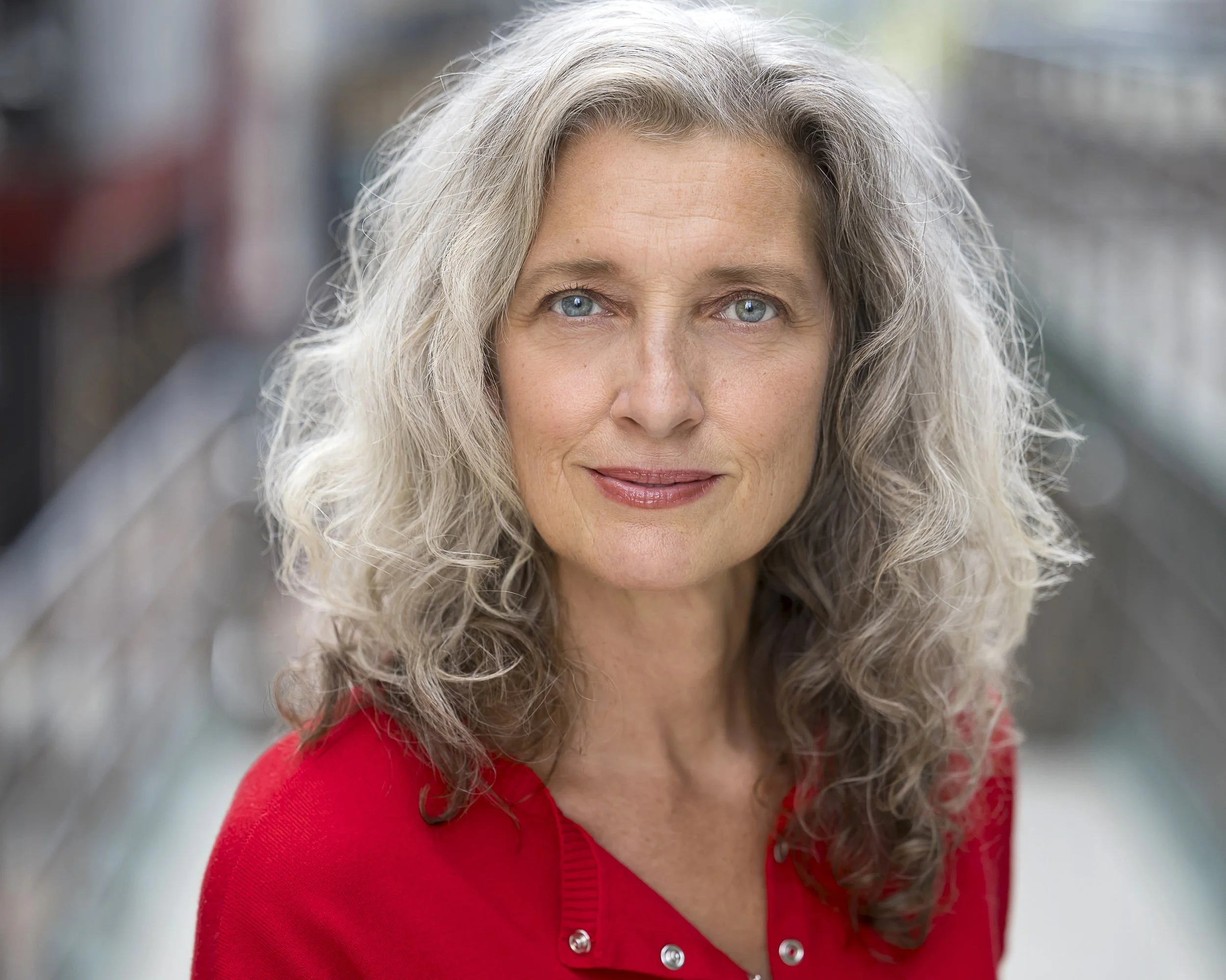 Close-up of an older woman with gray curly hair and blue eyes, wearing a red top with buttons, outdoors in a blurred background.