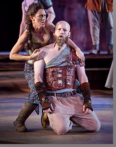 A man kneeling on stage with a woman standing behind him, his hands on his knees, in a theatrical performance setting.