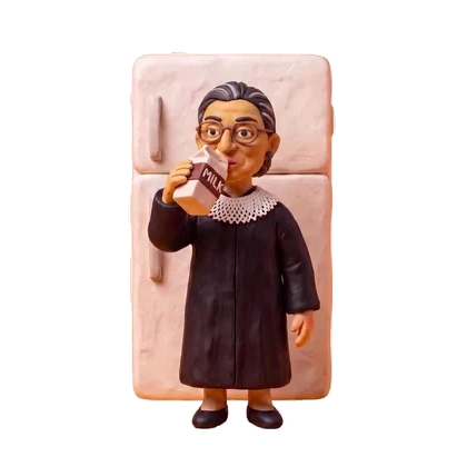 Figurine of Ruth Bader Ginsburg with glasses, gray hair, black robe, and white collar, standing in front of a pink refrigerator, drinking milk from a carton.