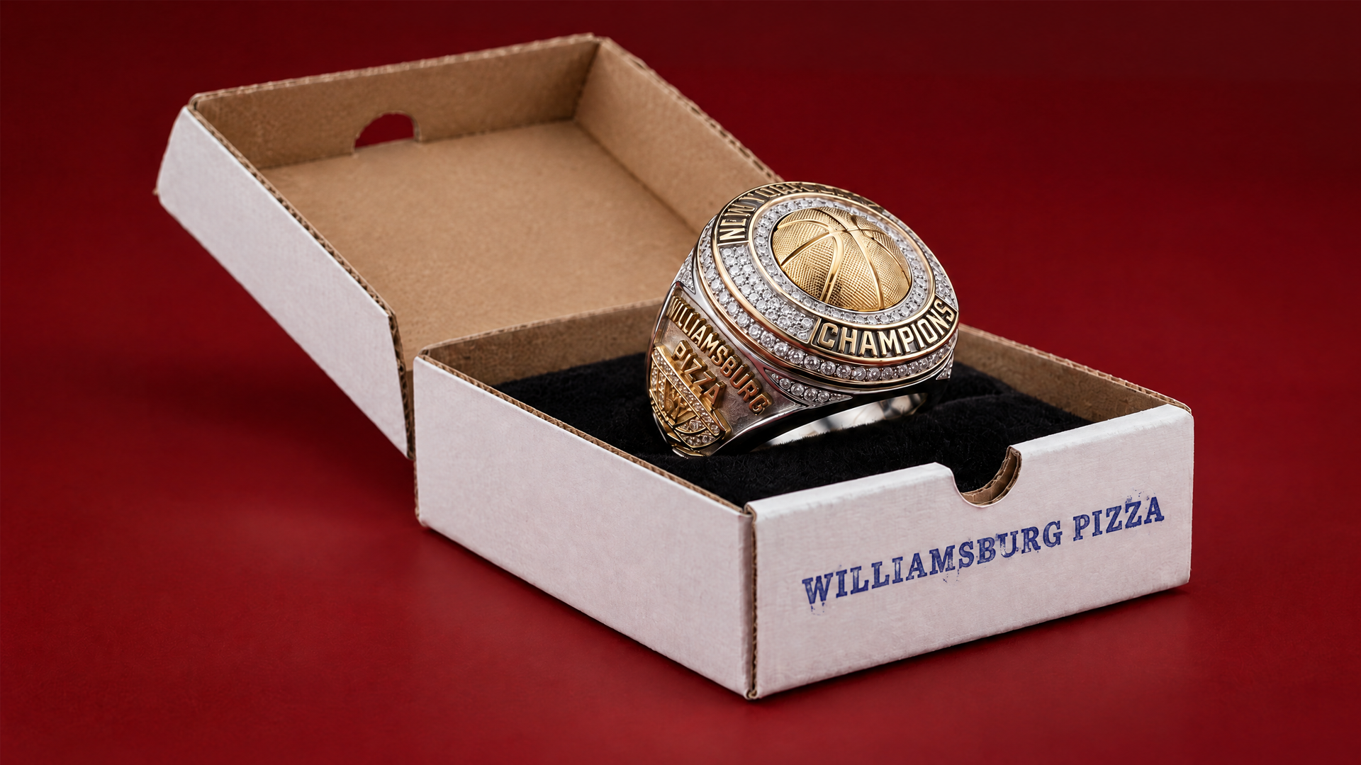 A championship basketball ring with gold and silver accents, engraved with 'WILLIAMSBURG PIZZA' and 'CHAMPIONS', inside a white cardboard box with black interior on a red surface.