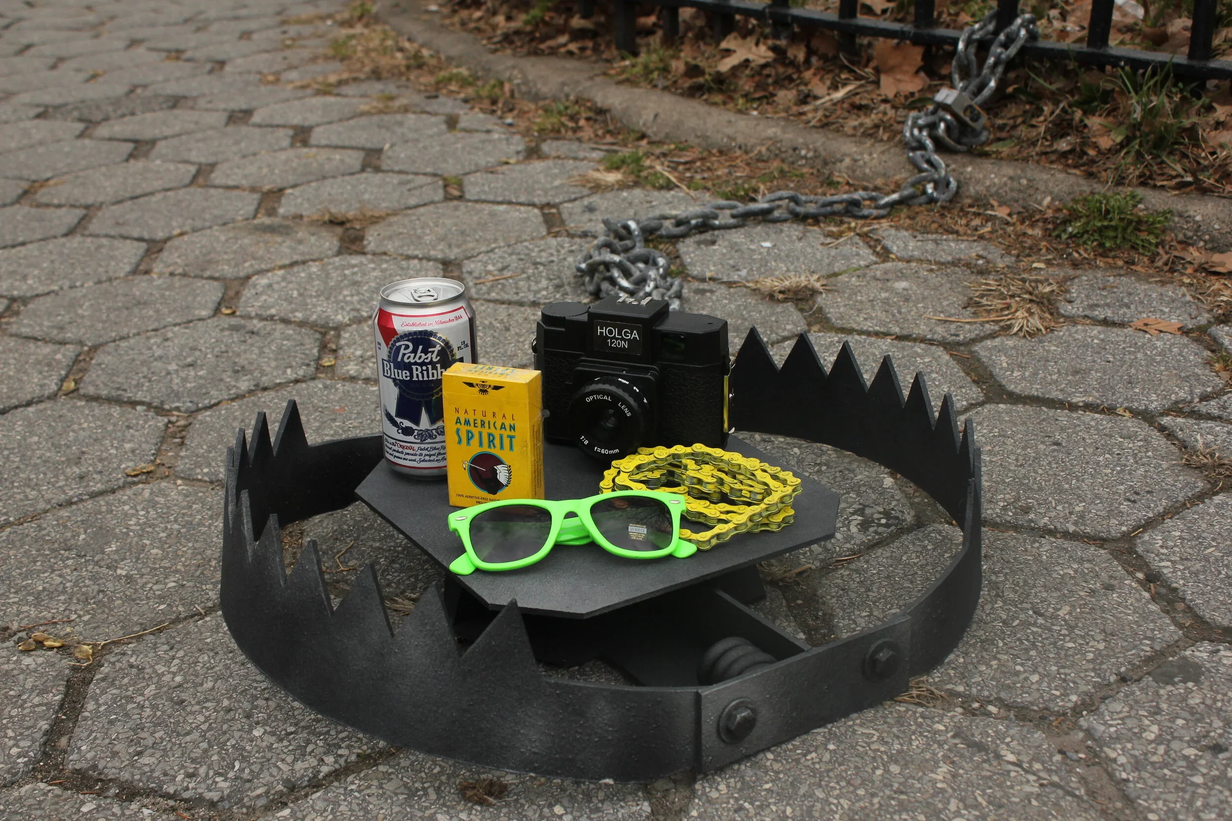Outdoor scene with a black fire pit table on a pavement, holding items including green sunglasses, a yellow bike chain, a can of Pabst Blue Ribbon beer, a yellow pack of American Spirit cigarettes, a black camera, and a small yellow object. A rusty chain and padlock are attached to a metal gate in the background.