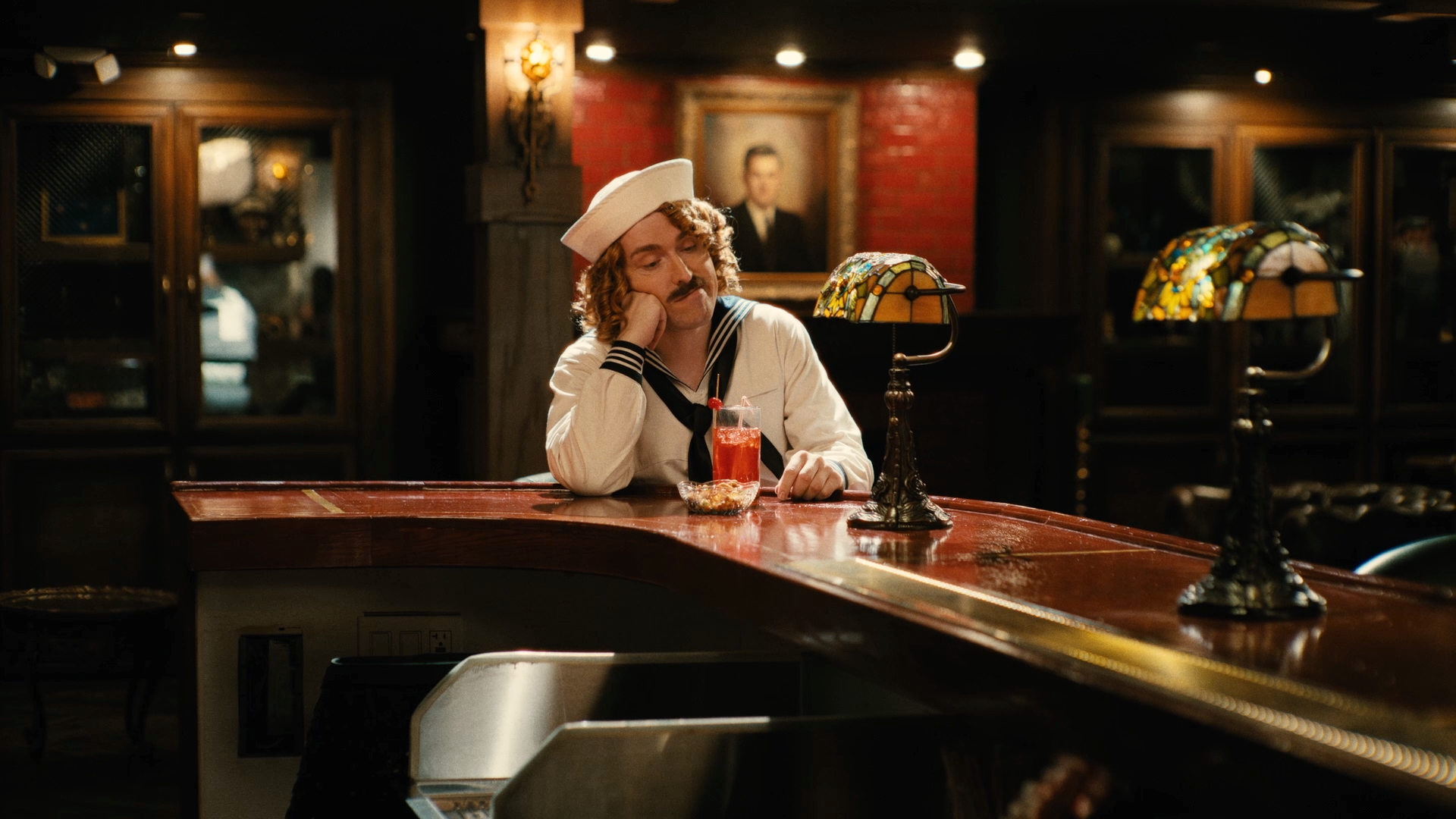 A man dressed as a sailor with a mustache, sitting alone at a bar counter with his head resting on his hand, and a red cocktail drink in front of him in a dimly lit bar.