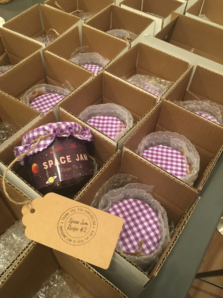 Boxes of small jars labeled 'Space Jam' with purple and white checkered cloth covers and bubble wrap inside, arranged in a cardboard box for sale or shipping.