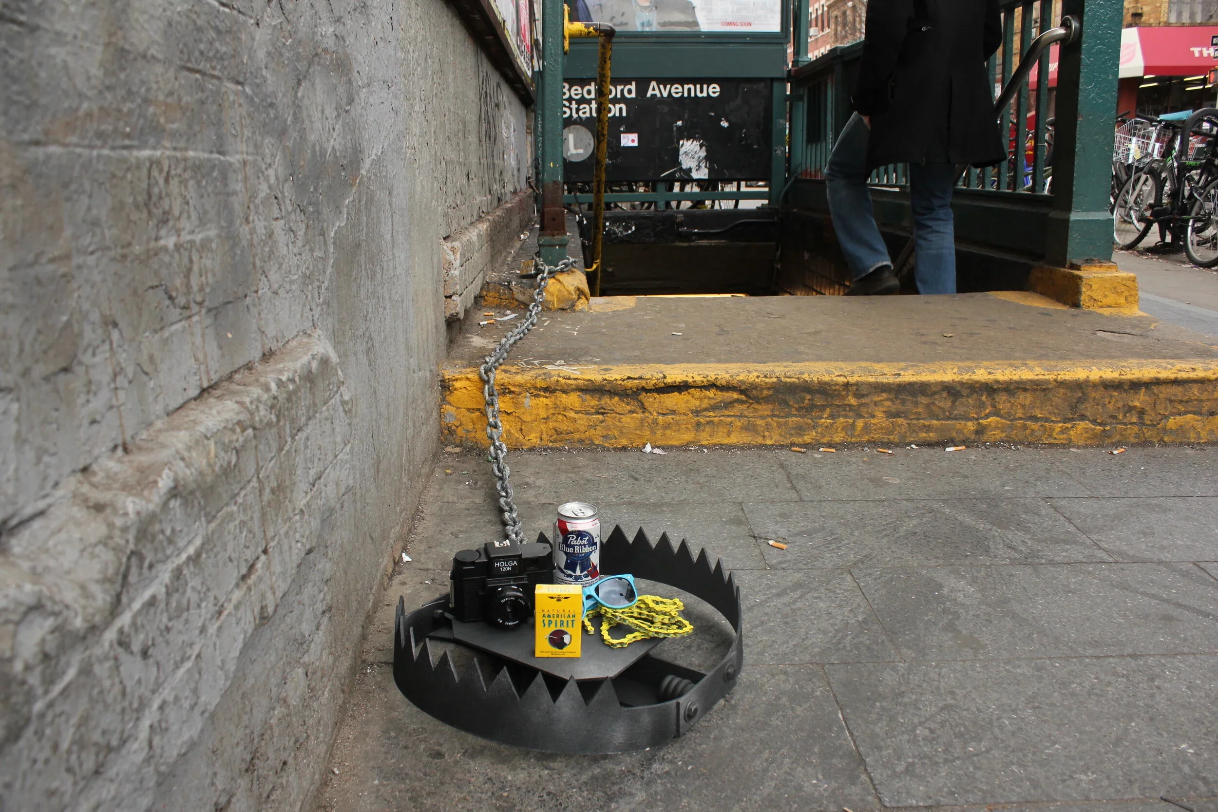 Underground subway entrance at Bedford Avenue station with warning devices in the foreground, including a saw, chains, and a can of Pabst Blue Ribbon beer on paved ground, and a person descending the stairs in the background.