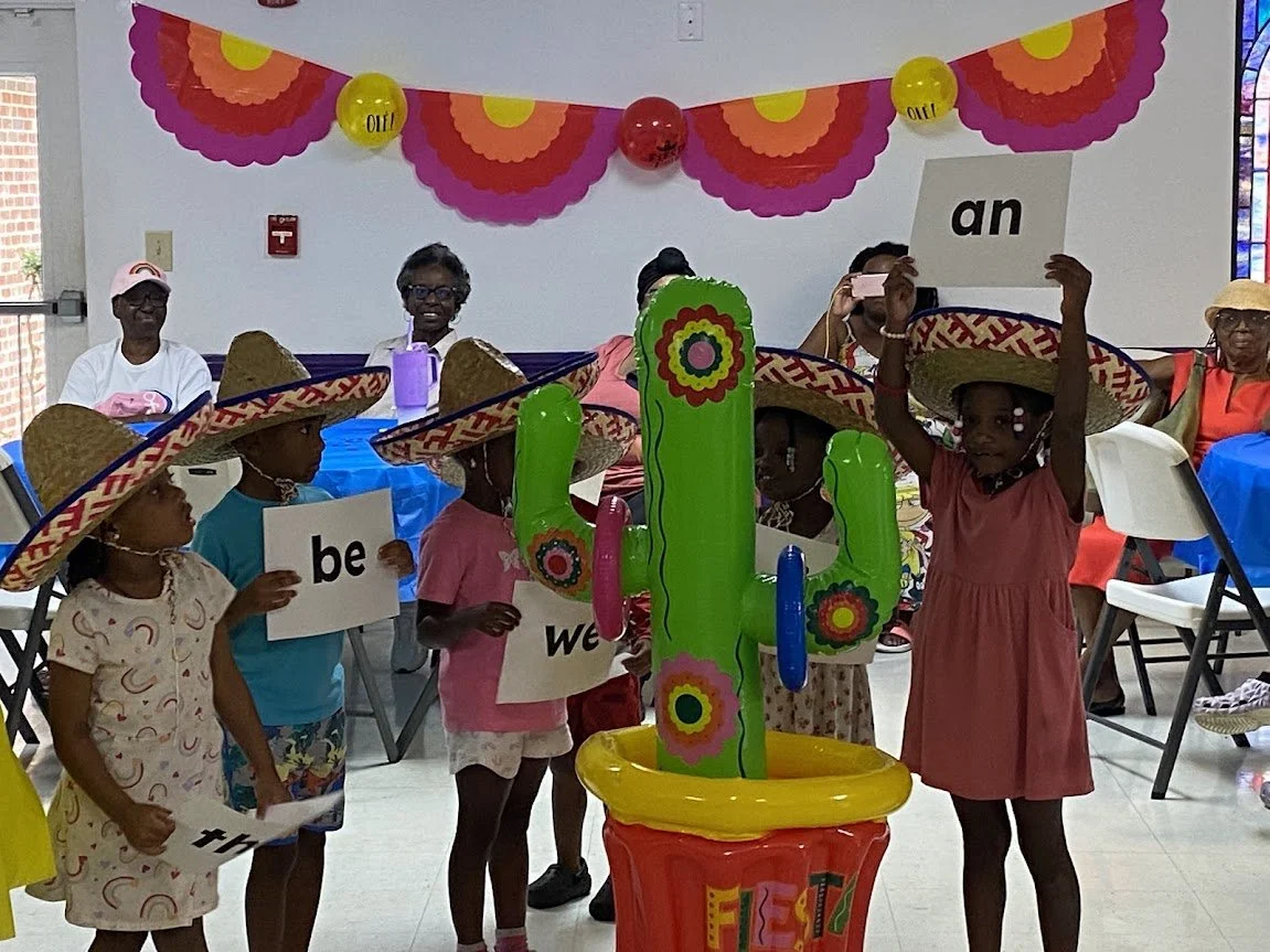 Children wearing sombreros holding signs with words, in a decorated room with balloons and a colorful banner.
