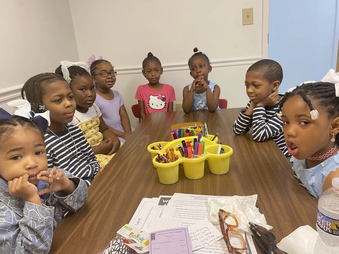 Children sitting around a table with crayons, paper, and glasses, in a classroom or meeting room.