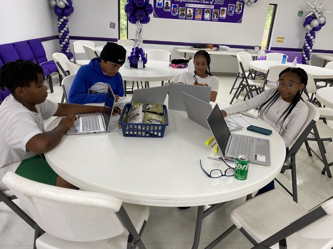 Four children sitting around a white circular table with laptops open in a decorated room, possibly for a graduation event, with purple and white balloons and banners on the wall.