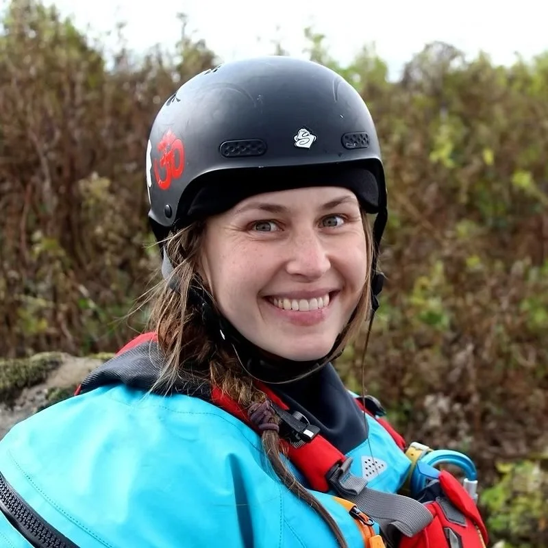 A smiling woman wearing a black helmet and outdoor gear, standing in front of foliage.