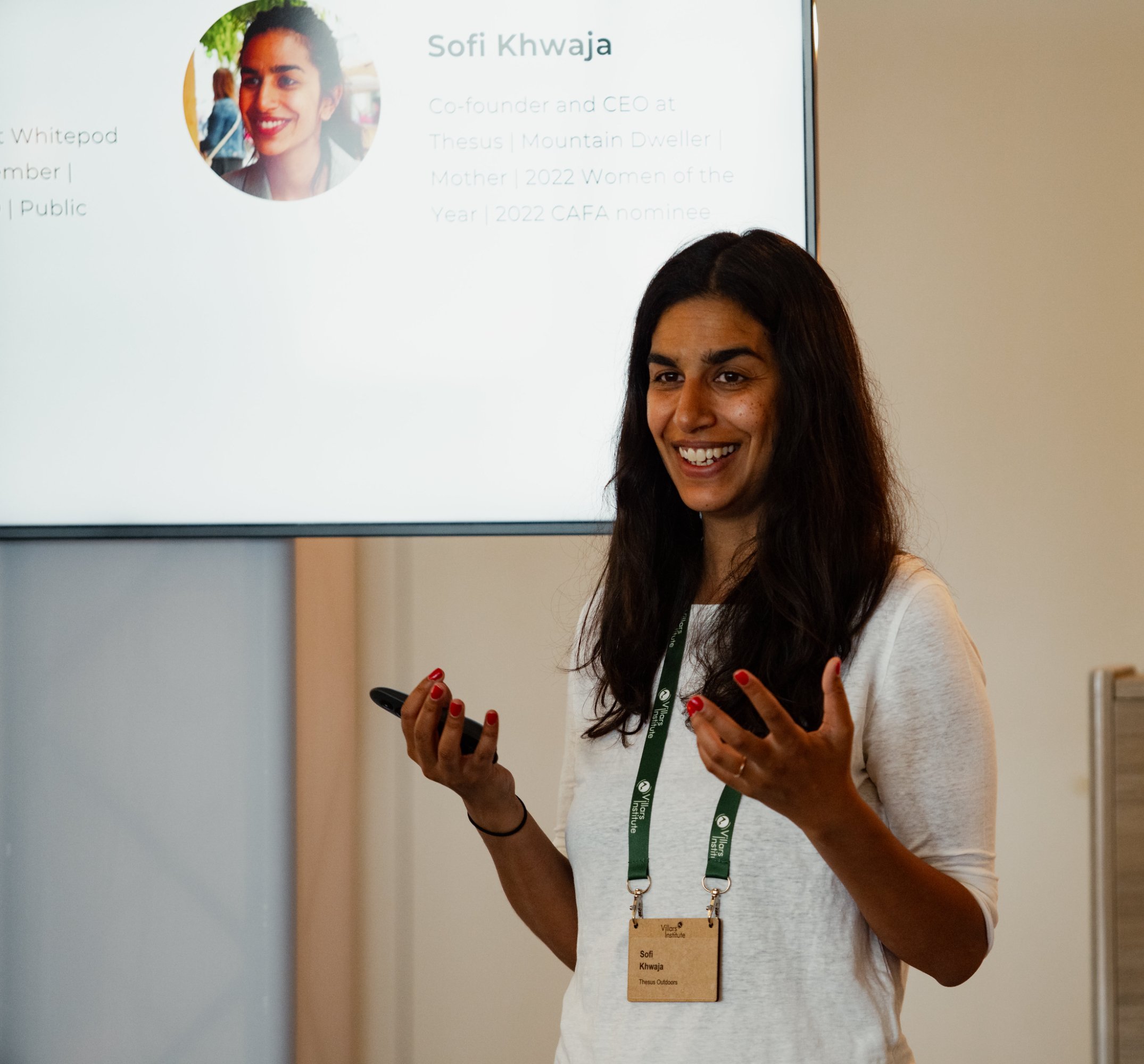 Woman with long dark hair, wearing a white shirt and green lanyard, giving a presentation in front of a slide with her photo and bio.