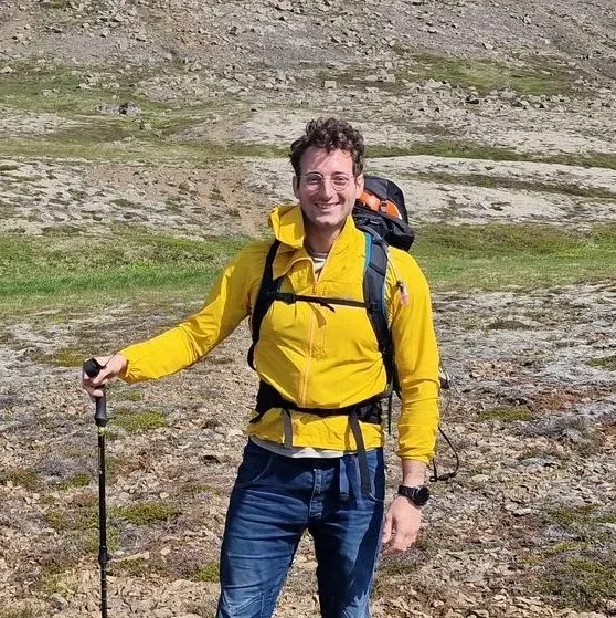 Man smiling outdoors in a yellow jacket, holding hiking stick, with backpack in a rocky, grassy landscape.
