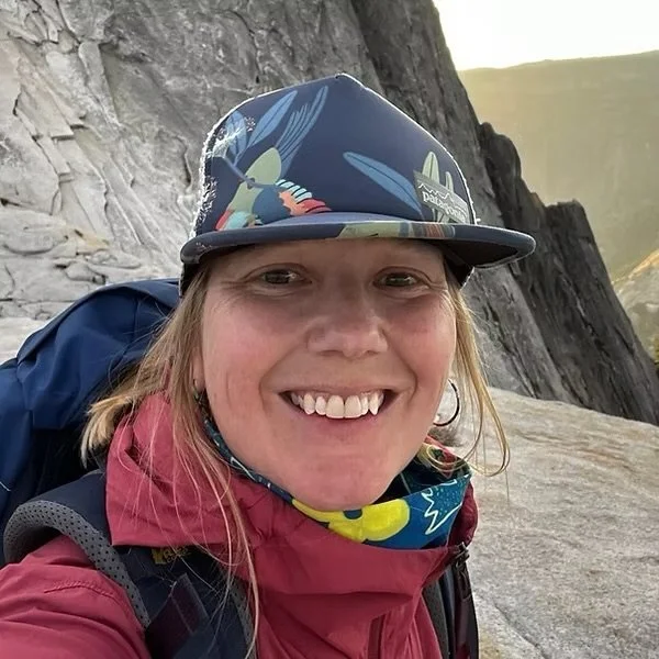 A woman smiling outdoors in a mountainous area, wearing a colorful cap with a shorter brim, a pink jacket, and a bandana around her neck, with a backpack on her shoulder.
