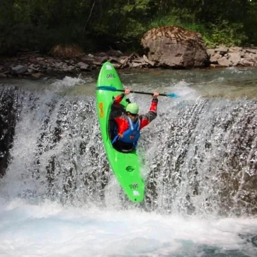 A person kayaking on a waterfall, wearing a helmet and a life jacket, with a paddling stick, surrounded by rocks and forest.