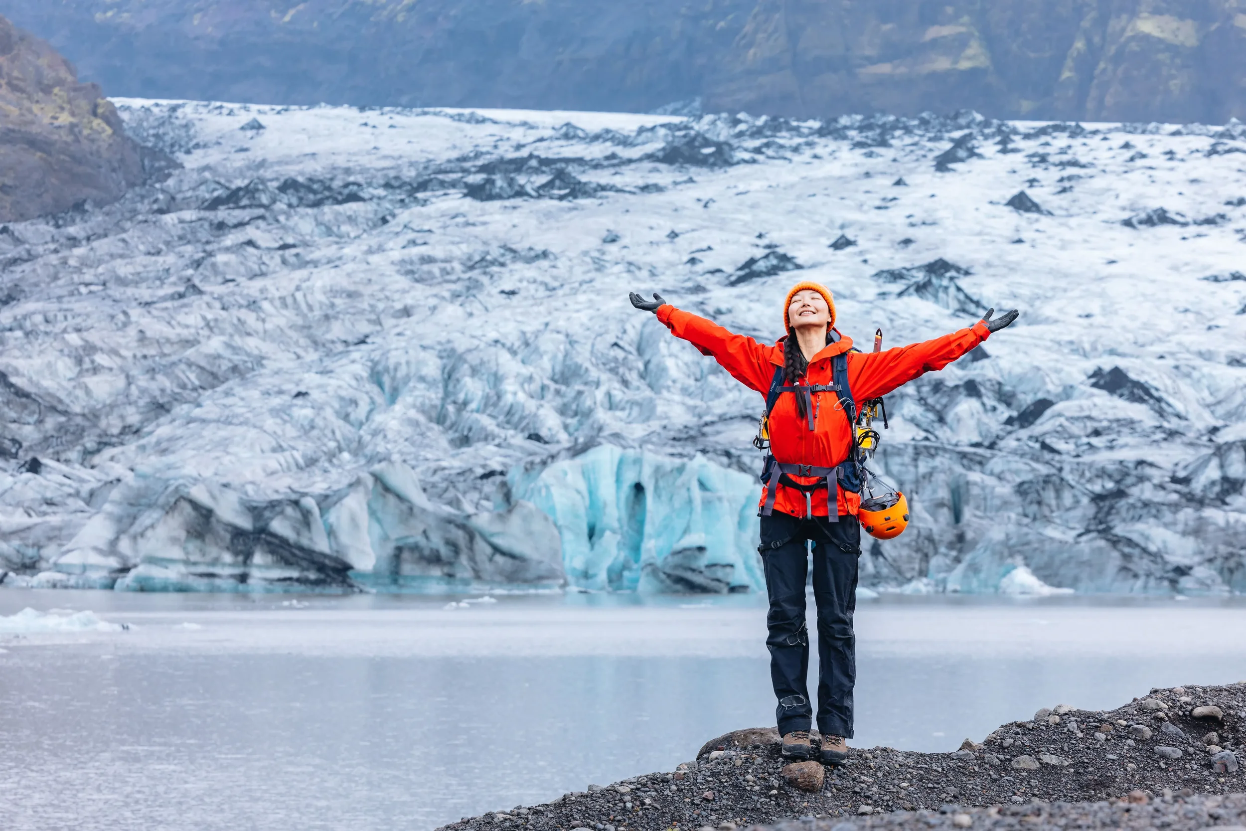 A woman in orange outdoor gear standing with arms outstretched in front of a glacier with a lake, smiling with her eyes closed.
