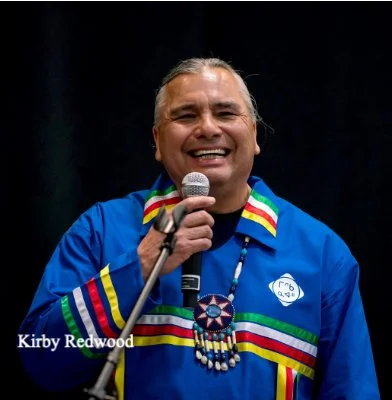 A man wearing a blue shirt with rainbow-colored stripes and a beaded necklace, smiling while holding a microphone.