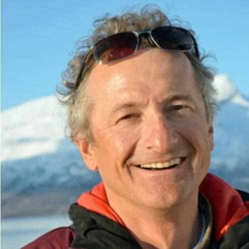A smiling man with sunglasses on his head, in outdoor gear, with mountains and a body of water in the background.