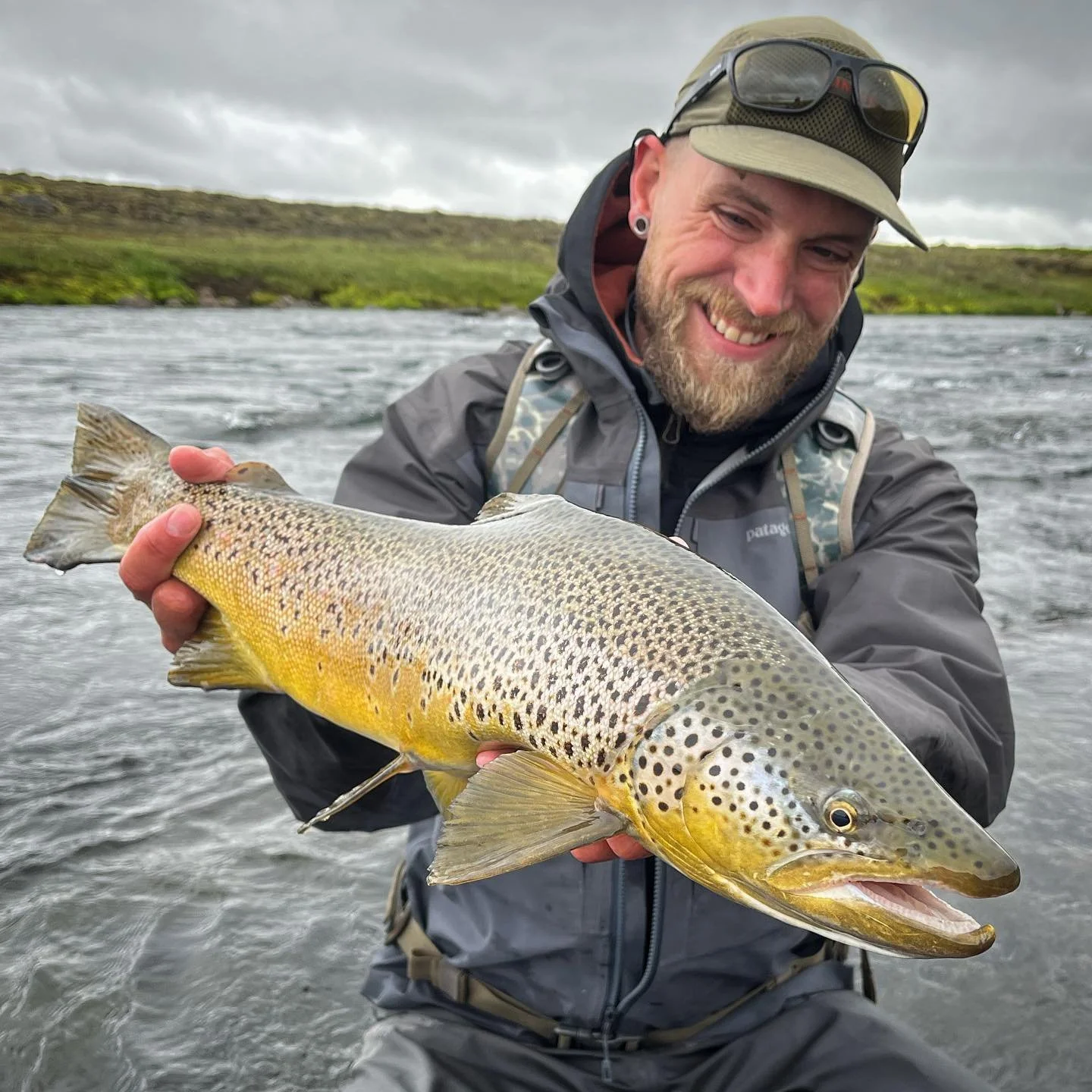 A man in outdoor gear holding a large fish by a river, with green hills and cloudy sky in the background.