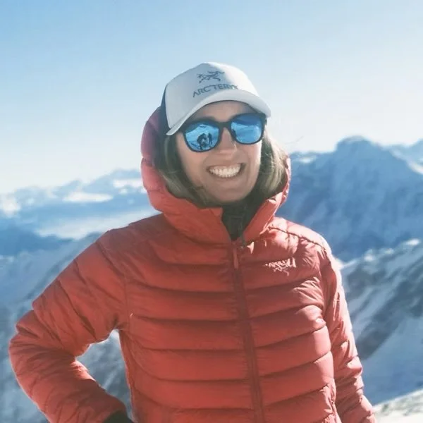 Smiling woman wearing sunglasses, a white hat, and a red jacket, with snowy mountains in the background.
