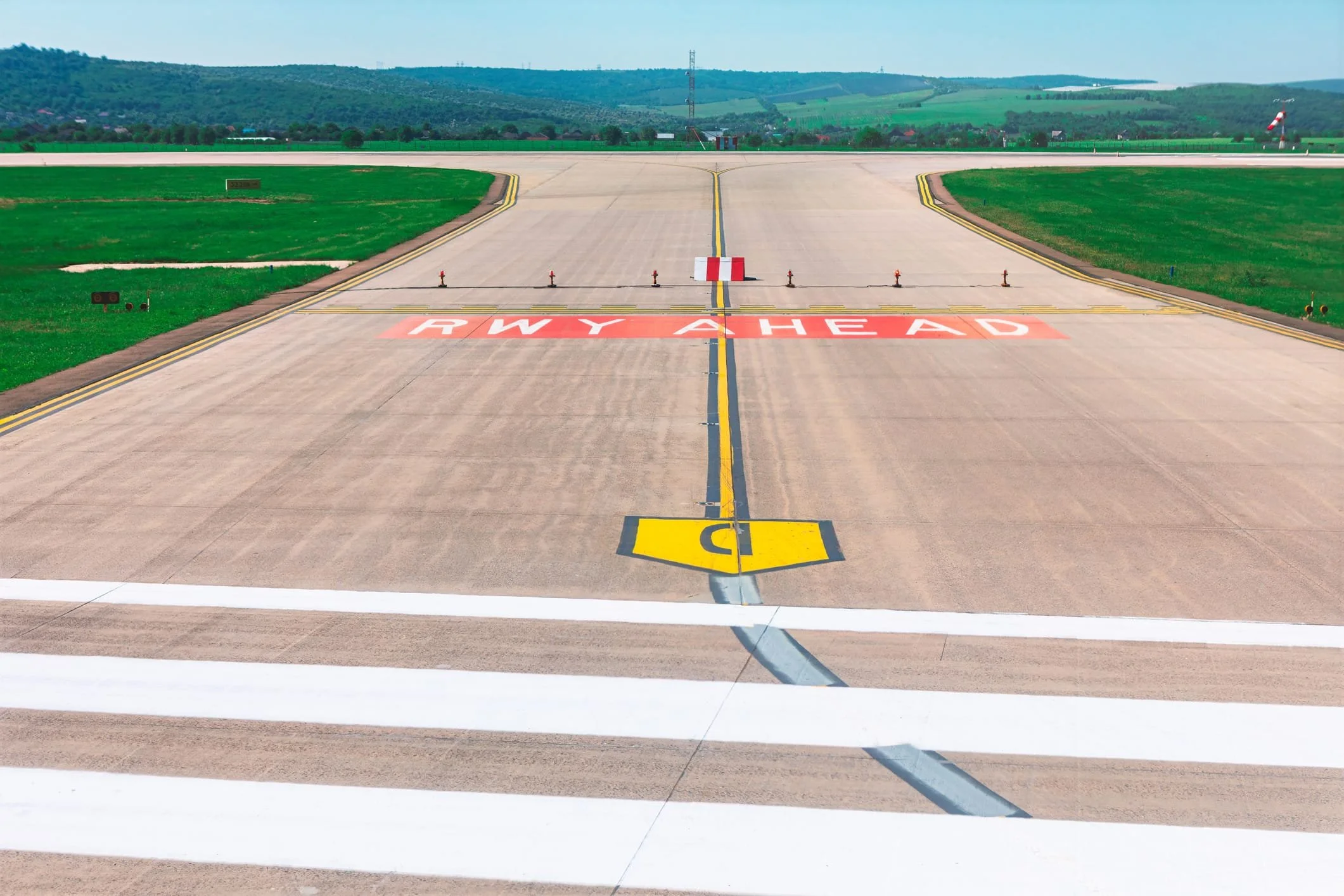 Runway at an airport with a red sign that says 'RWAY AHEAD' and a yellow arrow marker, surrounded by green fields and distant hills.