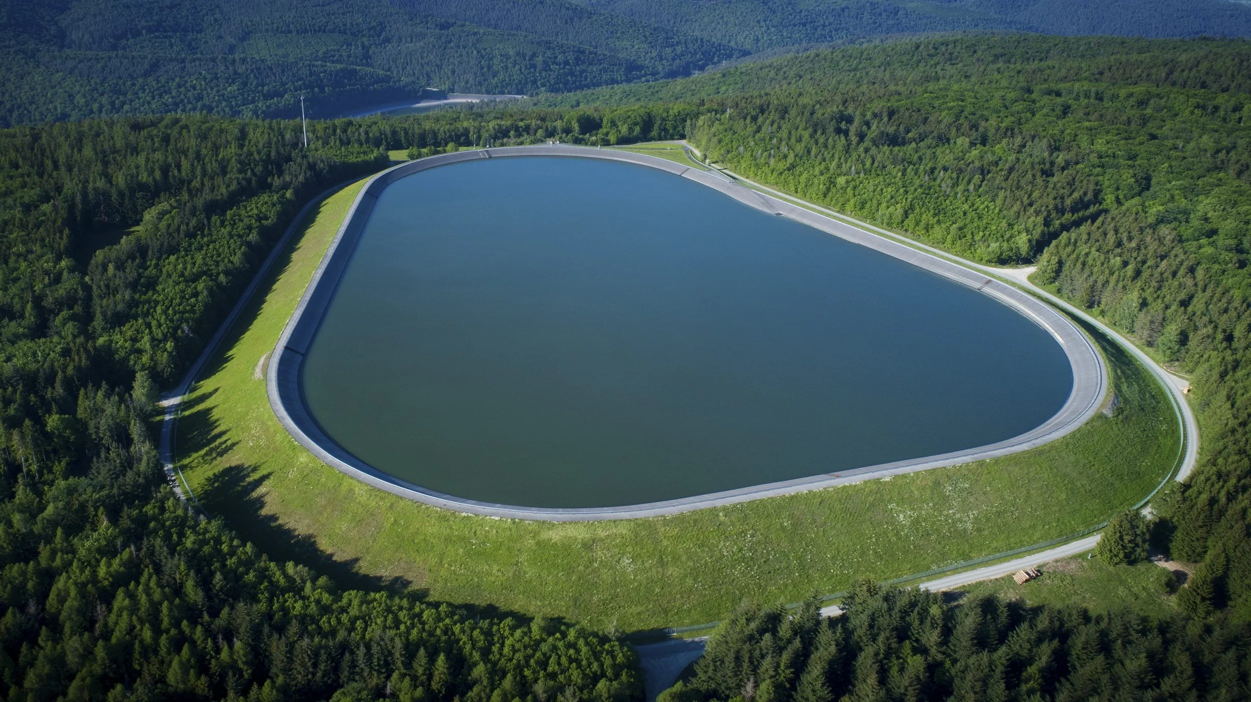 Aerial view of a large, curved dam holding back a body of water, surrounded by dense green forest in a mountainous area.
