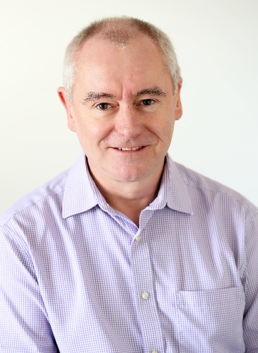 A middle-aged man with short gray hair, wearing a light purple checkered button-up shirt, smiling at the camera against a plain light background.