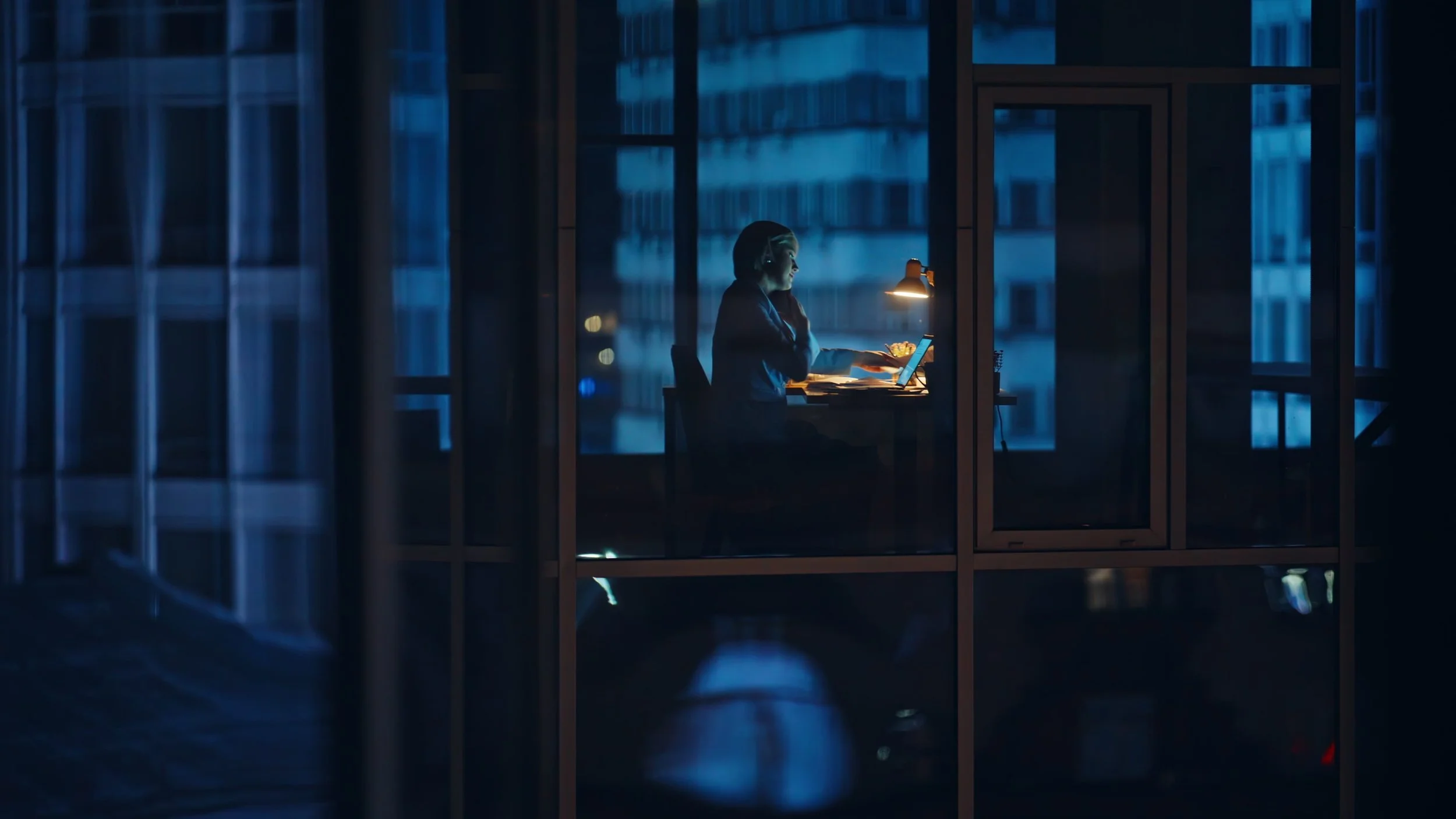A person working alone at a desk on a laptop in a glass-walled office at night, illuminated by a desk lamp, with a cityscape visible through the windows.