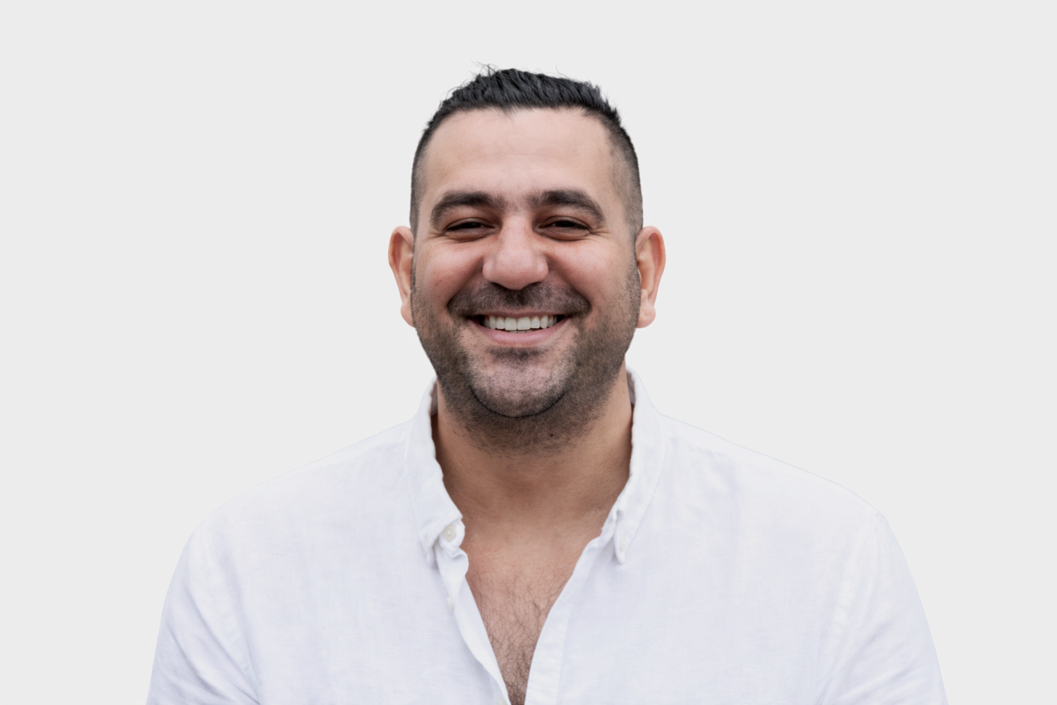 A man with dark hair and a beard smiling, wearing a white shirt, standing in front of green foliage.