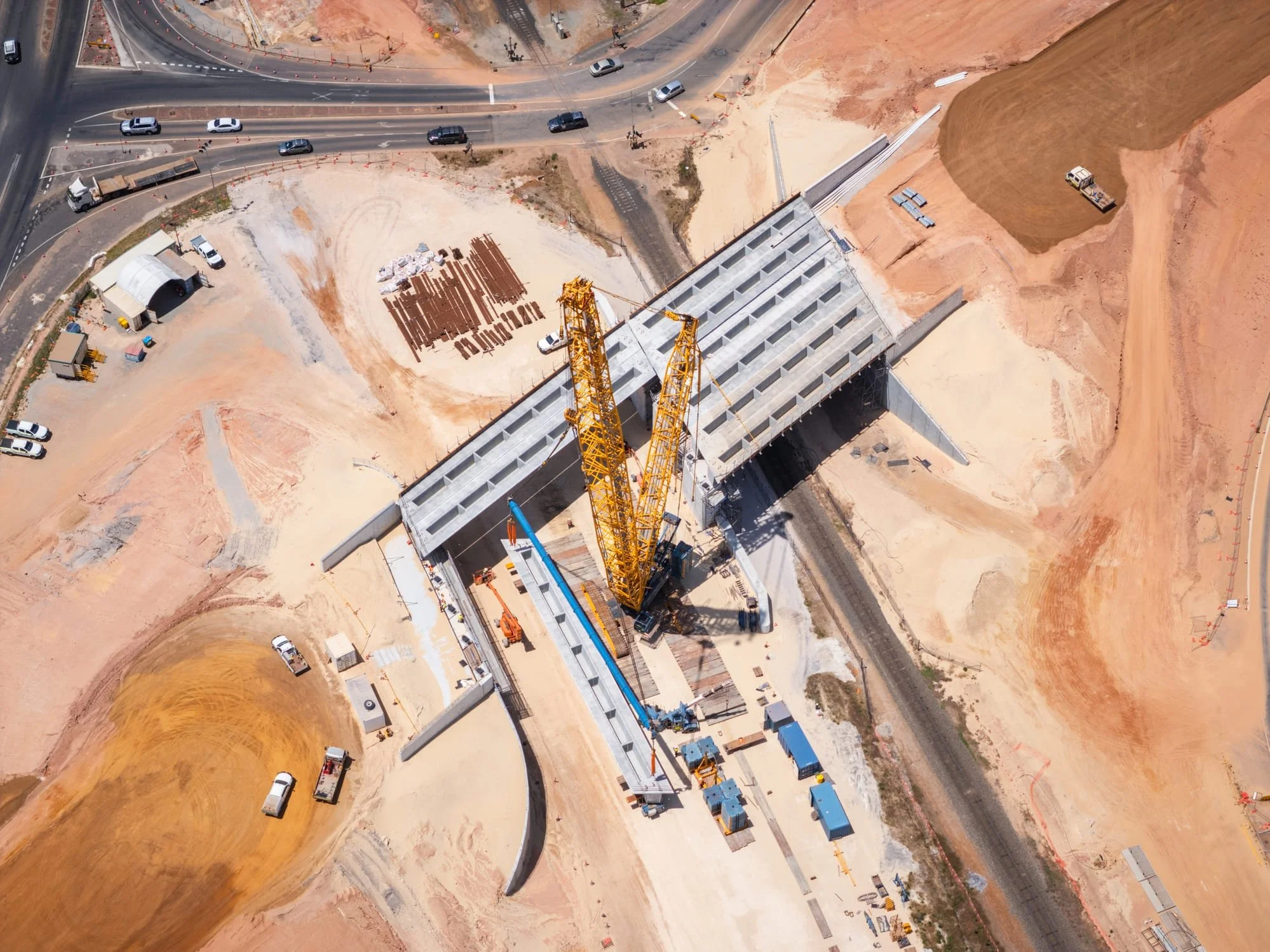 Aerial view of a highway construction site with a large yellow tower crane, road infrastructure, and vehicles. Extensive dirt and earth-moving areas surround the construction site.